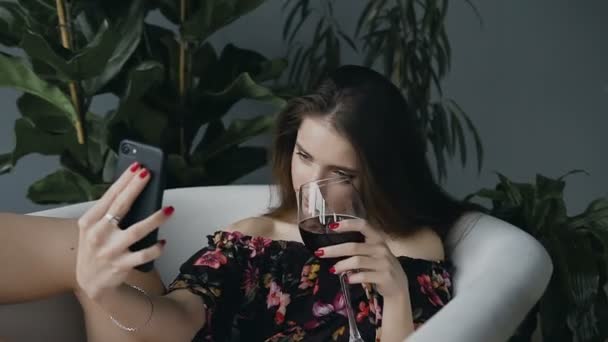 Una hermosa chica con una copa de vino tinto se relaja en un baño de leche caliente con brotes de flores fragantes y hace su foto selfie desde el teléfono — Vídeos de Stock