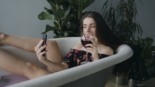 En söt tjej med långt hår avslappnad i en bubbelpool med mjölk och med färgglada doftande blommor knoppar, hon dricker gott rött vin och har kul chattar via internet nätverket via telefon — Stockvideo