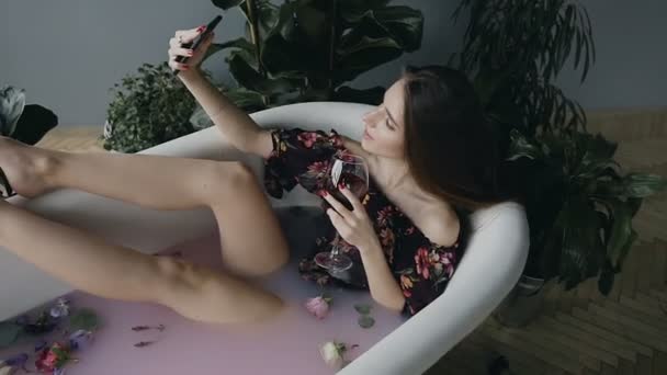 Красива дівчина робить селфі на телефоні в ванні, вона розслабляється в теплій молочній ванні з ароматними квітами і п'є червоне вино з келиха — стокове відео