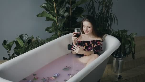 Una ragazza sexy con un bicchiere di vino rosso si siede e si rilassa in un bagno di latte caldo con boccioli di fiori profumati e utilizza il telefono su Internet. Giovane donna fa selfie foto con il suo telefono in bagno — Video Stock