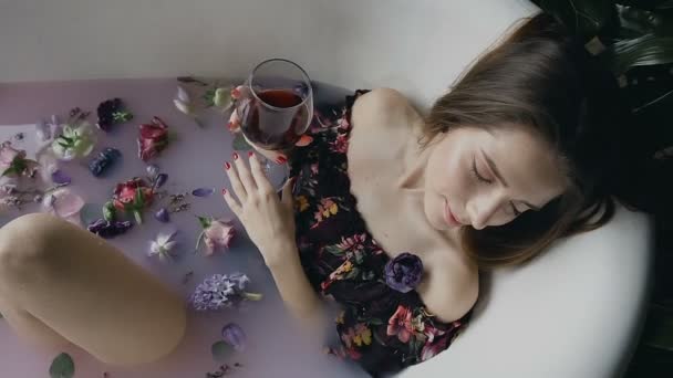 Sexig tjej är avslappnande i ett varm mjölk bad med väldoftande blomknoppar och dricka läckra rödvin från glaset. Aromaterapi, kroppsvård. Hälsosam livsstilskoncept — Stockvideo
