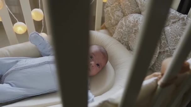 In der Krippe liegender neugeborener Junge blickt in die Kamera. niedliches kleines Baby, das zu Hause im Kinderbett liegt. Junge — Stockvideo