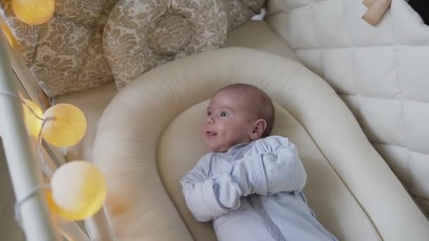 En nyfödd baby pojke i blå baby reglagen ljuger i spjälsängen hemma i sovrummet. Baby pojke försöker somna i barnsäng. Två månaders pojke — Stockvideo