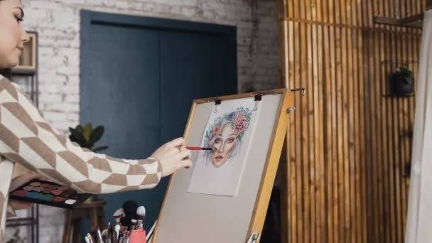 O artista em maquiagem e beleza cria uma bela tabela de maquiagem facial usando papel aquarela, sombras, batons e blushes usando um cavalete no estúdio. Jovem mulher em um estúdio brilhante desenha uma tabela facial — Vídeo de Stock