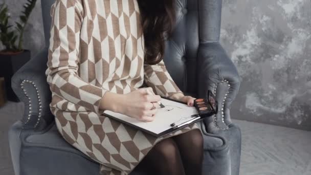 Face chart. Belle femme brune assise sur le fauteuil doux à la maison et dessine un tableau à l'aide du pinceau pour les cosmétiques et la palette d'ombres sur le papier aquarelle — Video