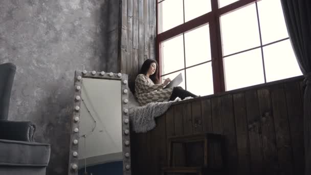 Artista femenina inspirada, en el dibujo del vestido cerca de la ventana. Chica morena atractiva está sentado en un alféizar de la ventana en el estudio de arte y dibujar en su cuaderno de bocetos — Vídeos de Stock