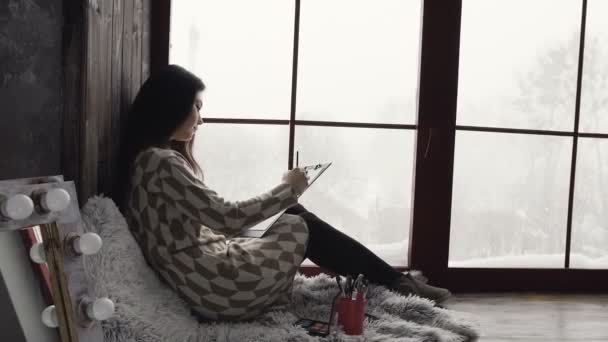 Jonge brunette vrouw zit op een vensterbank in de buurt van venster en tekening in haar schetsboek. Geïnspireerd artieste in jurk tekening in de buurt van het venster — Stockvideo