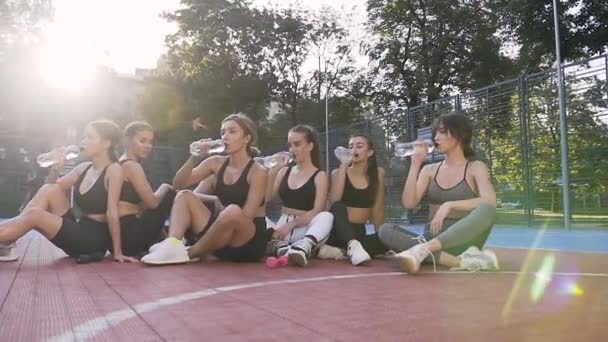 Γυμναστική γυναικεία νεαρή ομάδα κάθεται στο γήπεδο του αθλητισμού πάτωμα και πόσιμο νερό μετά την προπόνηση φυσικής κατάστασης — Αρχείο Βίντεο