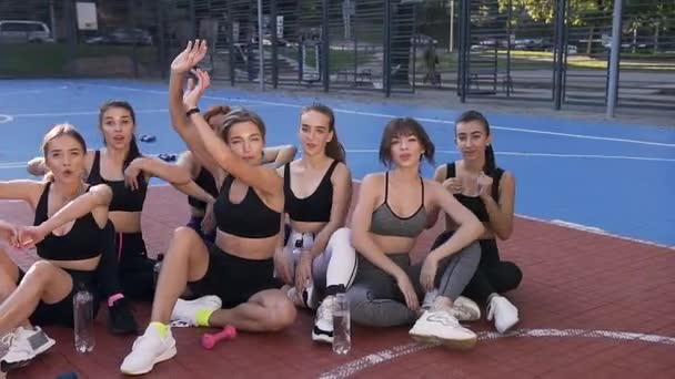 フィットネス女子チームが屋外競技場の床に座り、スポーツトレーニングの後に手を振って — ストック動画