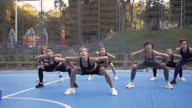 Female fitness team with instructor doing squat exercises on the sportground in the park in daytime