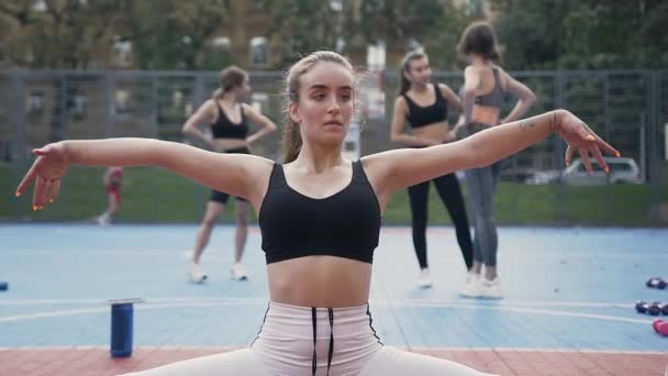 Giovane bella donna in abbigliamento sportivo seduto a terra e fare spago sullo sfondo del suo team di ragazze in forma fisica — Video Stock