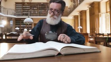 Wise male pensioner with beard in glasses sitting in the library and flipping through book pages,reading using magnifying glass