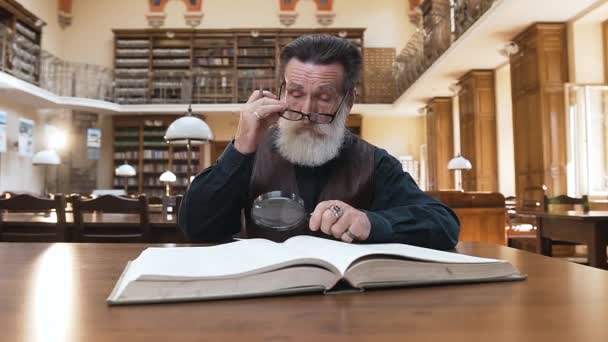 Viejo barbudo inteligente quitándose las gafas y frotándose los ojos durante su trabajo con un libro en la biblioteca — Vídeos de Stock