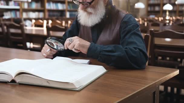 Vista lateral del atractivo anciano barbudo en gafas que se sienta en la biblioteca y trabaja con libro usando lupa — Vídeos de Stock