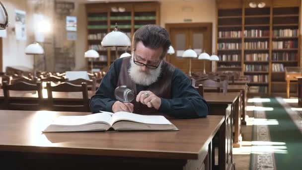 Vista frontal do homem velho concentrado inteligente em óculos que gravam notas importantes do livro, sentado na biblioteca — Vídeo de Stock