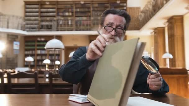 Attractive bearded senior man sitting at the library table ,taking off his glasses and closing book posing on camera — Stock videók