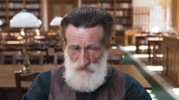 Attractive portrait of good-looking old bearded man which taking off his glasses and rubing the eyes after reading book and looking at camera — Stockvideo