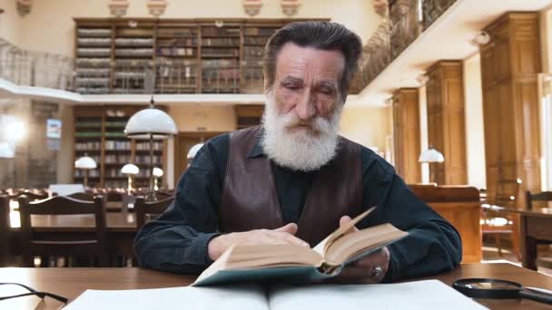 Крупним планом зосереджений старий бородатий чоловік, який перевертається через сторінки книг, сидячи за бібліотечним столом — стокове відео
