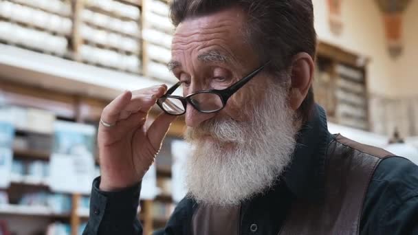 Vista lateral de un hombre barbudo mayor serio con anteojos que lee en la biblioteca — Vídeos de Stock