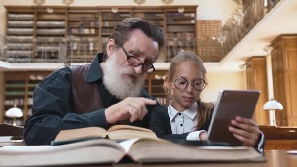 Snygg fläderskägg farfar sitter vid bordet i biblioteket med sin vackra sondotter och jämföra tryckta bok med ebook — Stockvideo