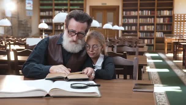 Gözlüklü yaşlı sakallı büyükbabanın ve kütüphanede oturup kitap okuyan güzel bir torunun güzel görüntüleri. — Stok video