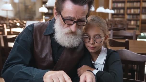 Ritratto attraente di nonno anziano piacevole con barba bene curata che legge il libro la sua nipote adolescente carina nella biblioteca — Video Stock