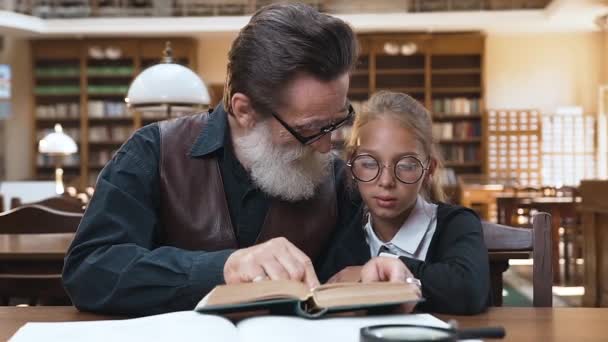 Vriendelijke senior opa met grijze baard zittend in de bibliotheek en leesboek met zijn schattige tiener kleindochter — Stockvideo