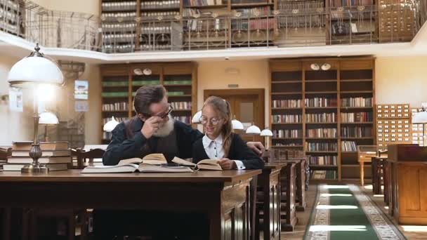 Homme âgé barbu concentré dans des lunettes assis à la table de la bibliothèque et écoutant comment lire sa jolie petite-fille adolescente — Video