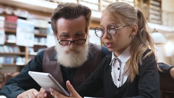 Viejo abuelo barbudo guapo con gafas hablando con su nieta adolescente sonriente y usando ebook en la biblioteca — Vídeos de Stock