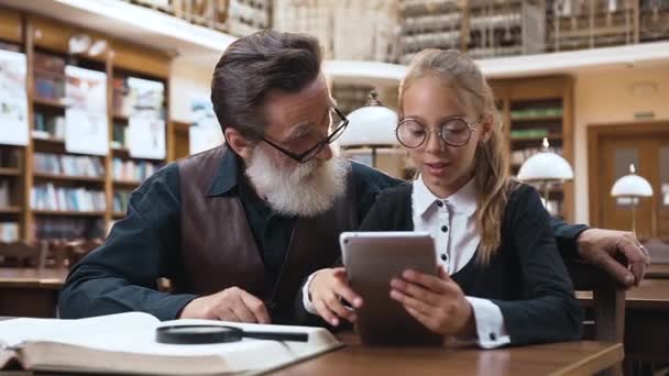 Gadis remaja cantik berkacamata membaca cerita dari buku elektronik sampai kakek janggut seniornya yang cerdas di perpustakaan — Stok Video