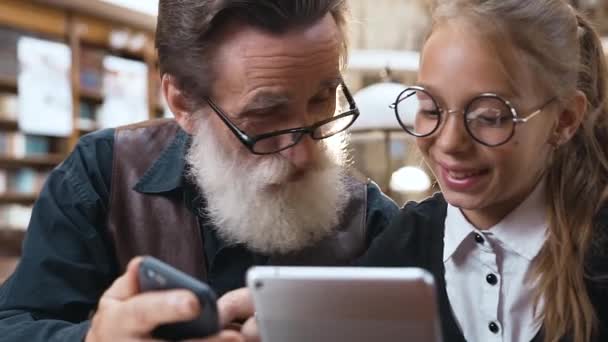 Intelligente senior man met gerimpeld gezicht en grijze baard die iets op zijn mobiel laat zien aan zijn lachende tienerkleindochter terwijl ze in de bibliotheek zitten — Stockvideo
