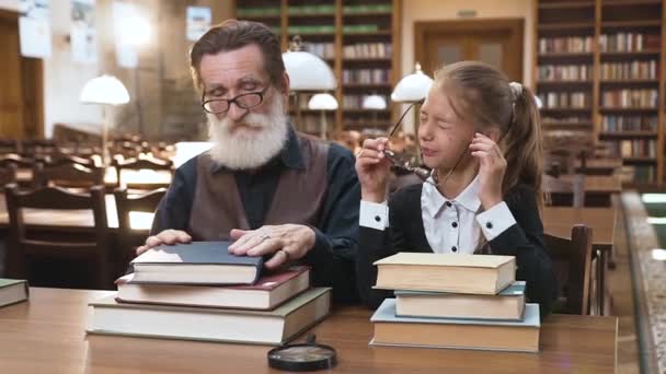 Moe, maar gelukkige man van 70 jaar met baard en zijn blonde, mooie kleindochter rustend op de boeken in de bibliotheek — Stockvideo