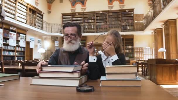 Привлекательный старый бородатый мужчина снимает очки со своей уставшей 12-летней внучкой и лежит на книгах после прочтения в библиотеке — стоковое видео