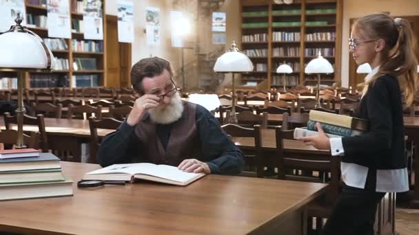 Starszy mężczyzna z brodą w okularach czyta książkę, podczas gdy jego blond wnuczka przynosi mu na stół trzy książki. — Wideo stockowe