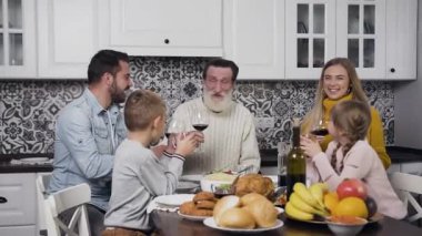 Güzel görünümlü mutlu bir aile, Şükran Günü kutlamaları sırasında ellerinde şarap ve meyve suyu kadehleriyle şenlik masasında oturuyor.