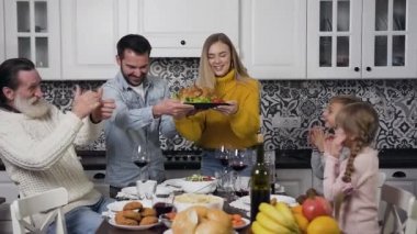 Sevimli yaşlı sakallı dede ve gülümseyen kız ve oğlan ellerini çırpıyor. Bu sırada neşeli ebeveynler Şükran Günü masasına geleneksel hindiyi koyuyorlar.