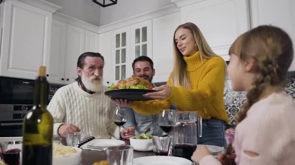 Krásná radostná mladá žena ve stylovém oblečení dávat do středu stolu chutné pečené krocana při oslavě Den díkůvzdání s celou její rodinou se starším dědečkem — Stock video