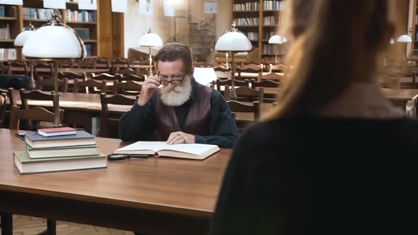Уважаемый старший бородатый мужчина в очках сидит за библиотечным столом, пока его милая внучка-подросток приносит ему толстые книги — стоковое видео