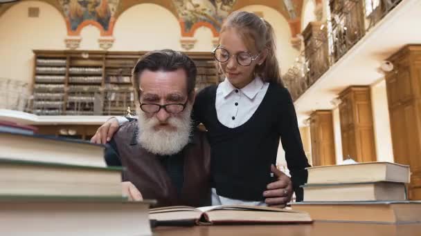 Viejo abuelo barbudo inteligente en gafas libro de lectura con su nieta adolescente lindo en la mesa de la biblioteca — Vídeos de Stock