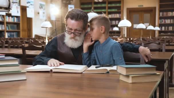 Bonito menino loiro sussurrando para seu velho avô experiente que está sentado na biblioteca — Vídeo de Stock