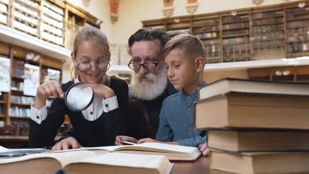 Feliz abuelo barbudo mayor leyendo emocionante libro a sus nietos adolescentes sonrientes en la biblioteca — Vídeos de Stock