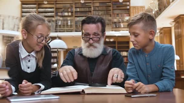 Розумний літній чоловік з доглянутою бородою сидить за бібліотечним столом і читає книгу зі своїми підлітками приємними онуками, використовуючи збільшувальне скло — стокове відео