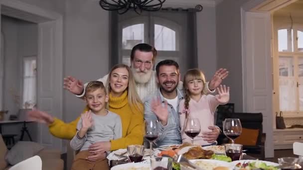 Сміються щасливі люди родичі сидять за святковим столом і дивляться на камеру з махаючими руками — стокове відео