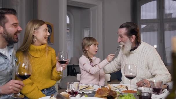 Retrato divertido de neta bonito que alimentando seu avô com um pedaço de frango na mesa de jantar, onde reunir toda a sua família — Vídeo de Stock