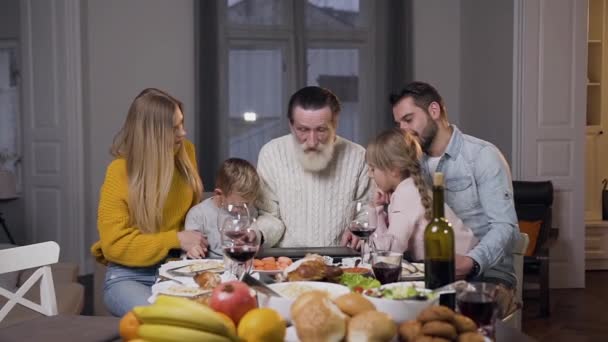 尊敬される祖父は夕食のテーブルの上にコンピュータを開き、彼の家族と一緒に写真を見て — ストック動画