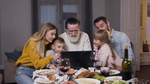 Jóképű, különböző generációs emberek ülnek a vacsoraasztalnál és vicces képeket néznek a laptopon. — Stock videók