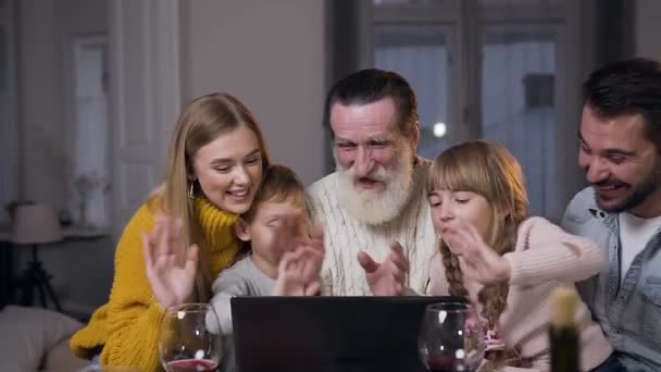 Alegre familia alegre con el abuelo sentado en la mesa festiva y tener video chat a través de la aplicación de mensajero en el ordenador y saludar a su interlocutor — Vídeo de stock