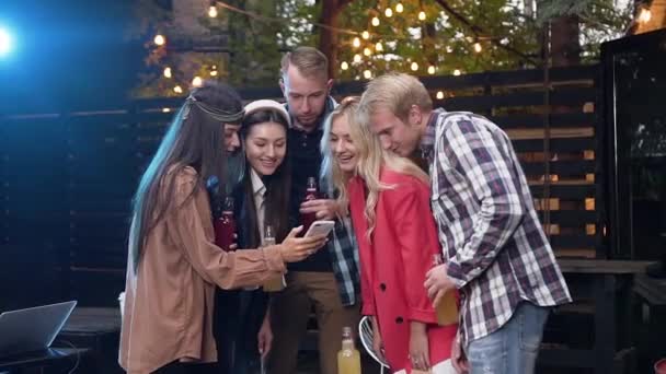 Радостные друзья-женщины и мужчины используют смартфон, разговаривая, держа бутылки с напитками во время празднования вечеринки. На открытом воздухе — стоковое видео