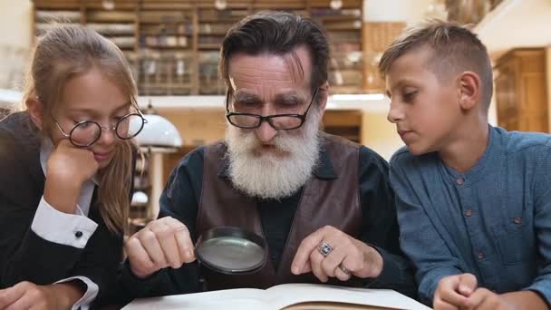 Intelligent old bearded granddad in glasses, reading book with magnifying glass with his interested teenage grandchildren in the library — ストック動画