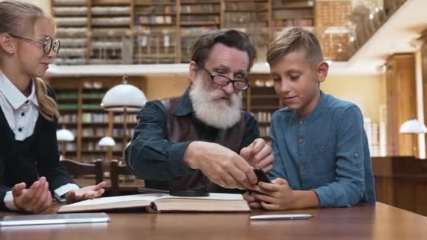 Vážný starý muž s upravenými vousy v brýlích odnáší telefon a ipad svým překvapeným vnoučatům a vysvětluje, proč je důležité číst knihy — Stock video